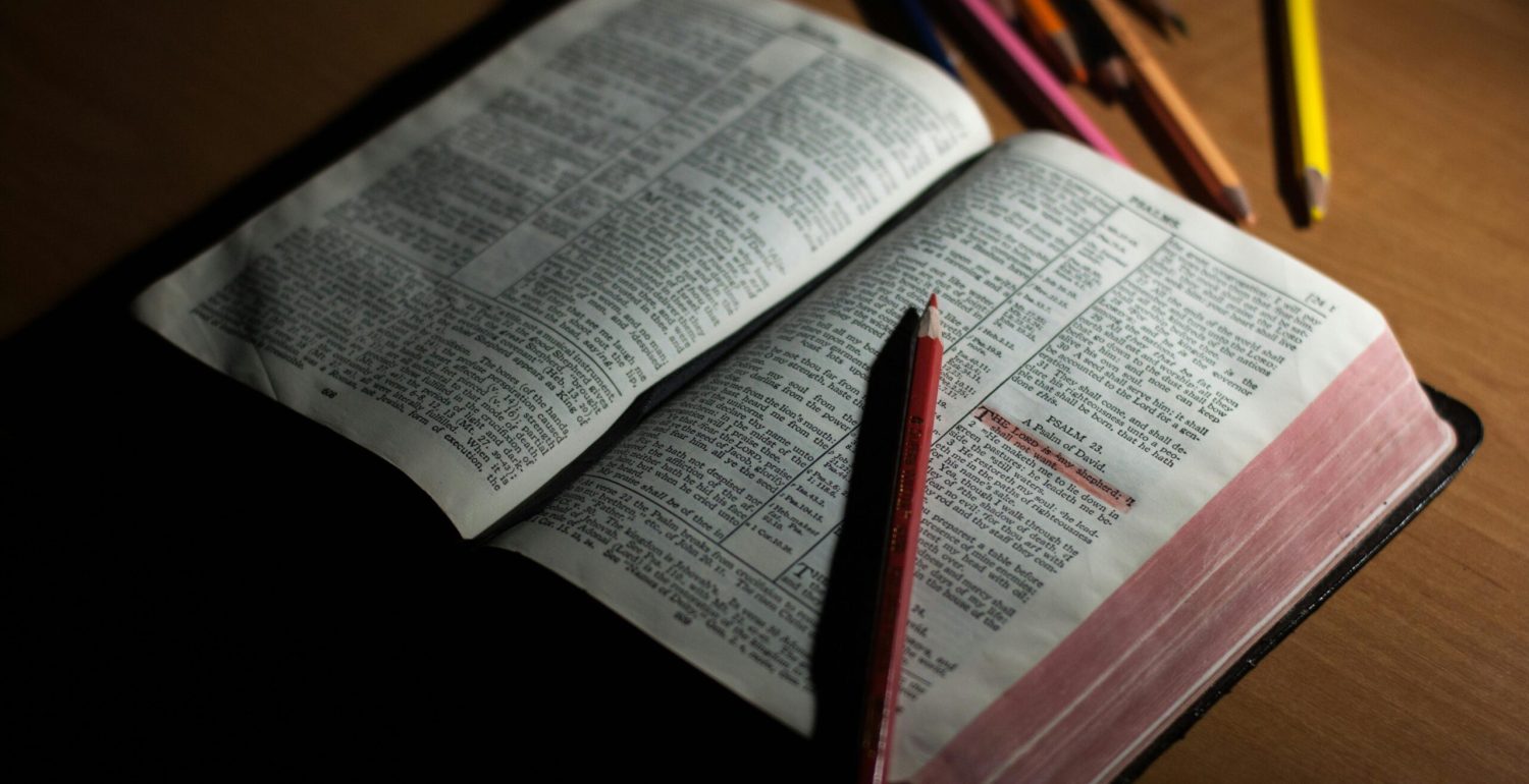 An open Bible on a wooden table accompanied by colored pencils, symbolizing study and spirituality.